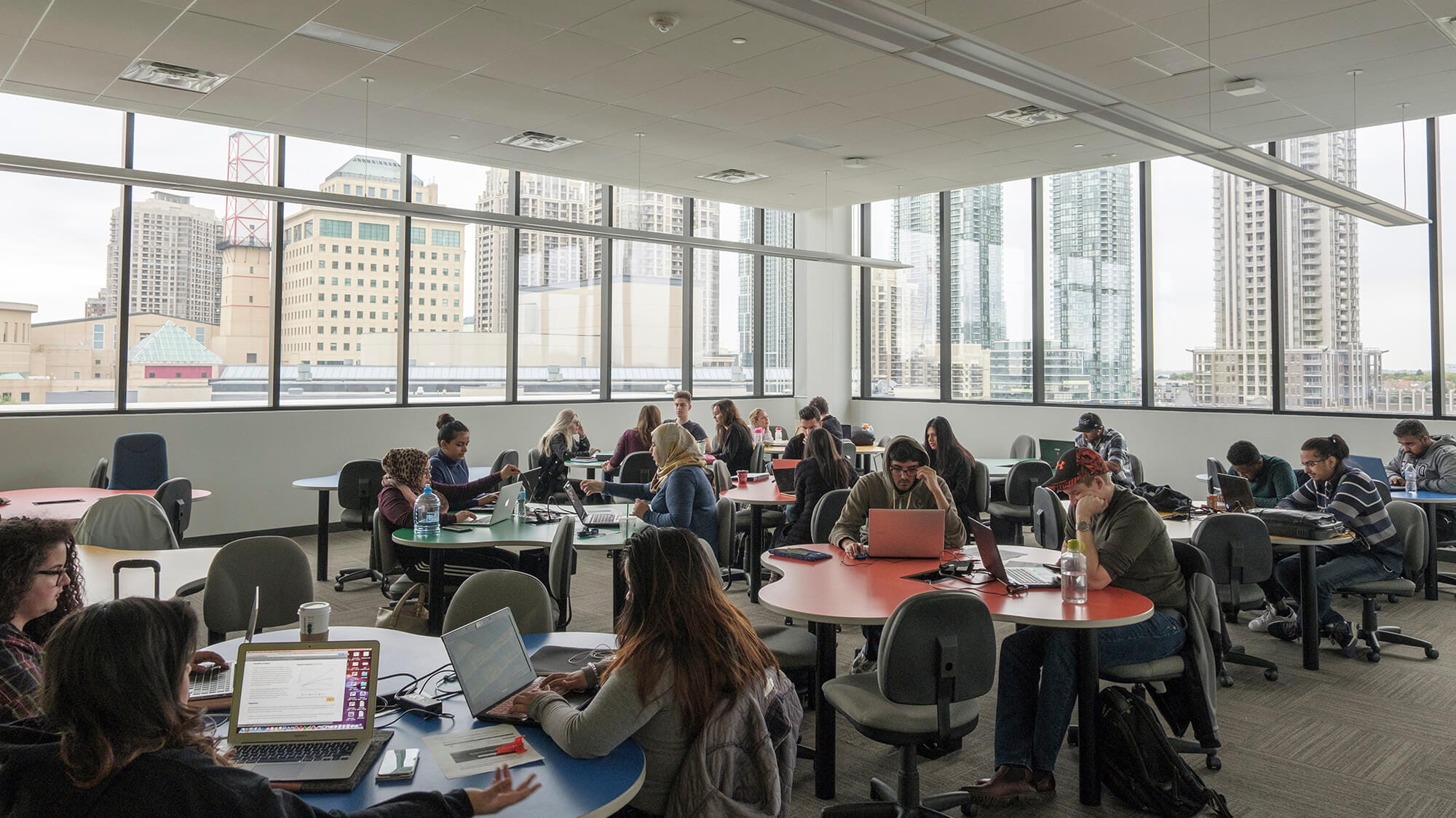 A bright room with lots of windows, filled with groups of students working at tables
