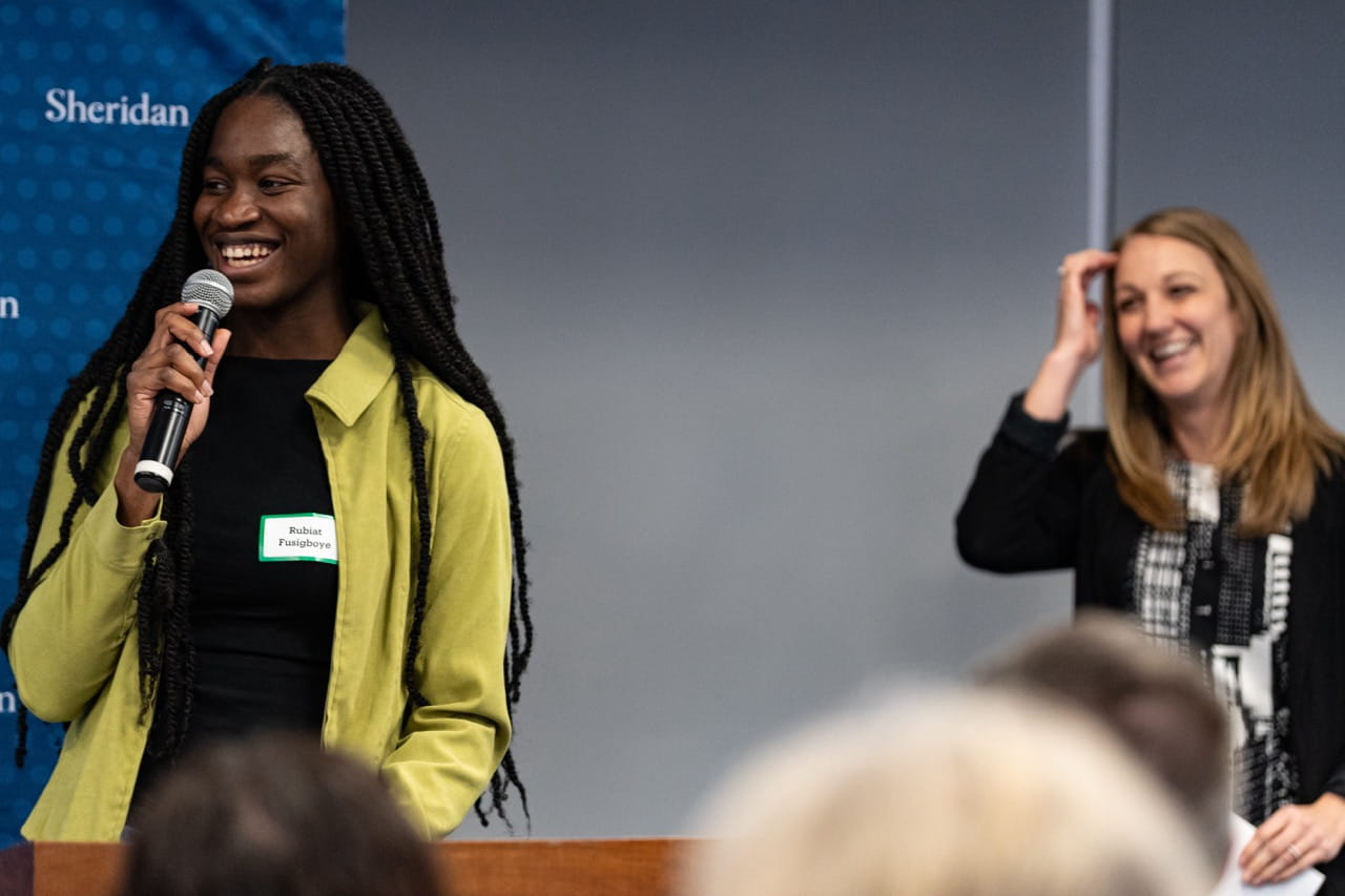 Rubiat Fusigboye speaks at the podium while Sheridan Experiential Learning Coordinator Jessica Angus smiles in the background