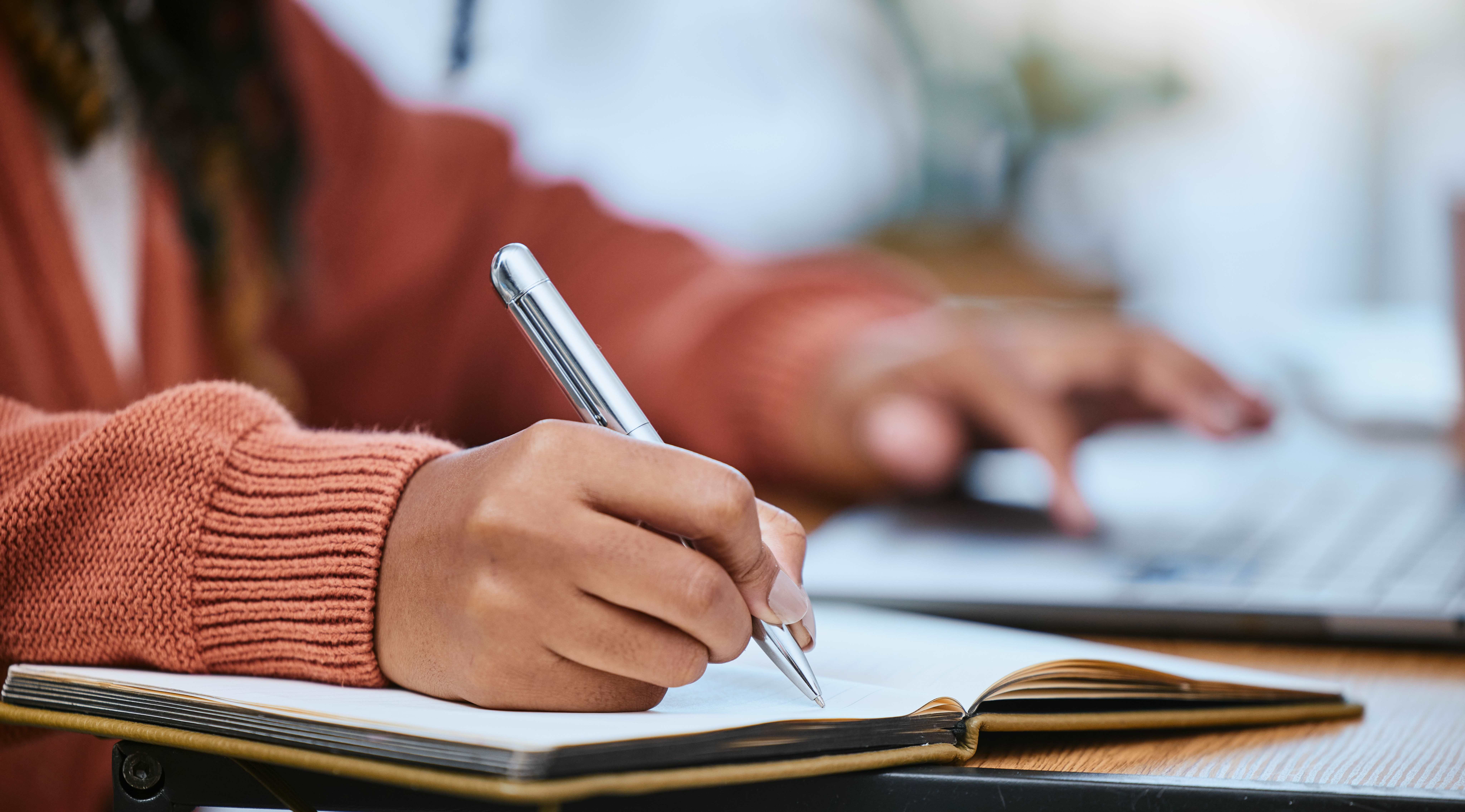 A person wearing an orange sweater holds a pen and writes in a notebook, with one hand one a laptop.