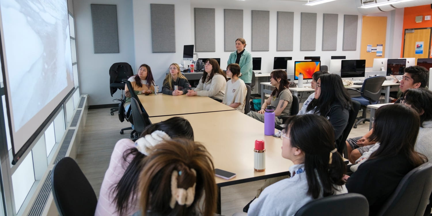 A group of students sits around a long table while an instructor teaches.